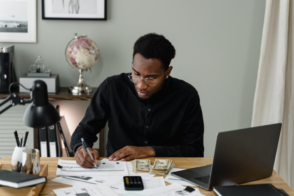 Young professional sitting at a desk, planning their budget with a laptop, paper, pen, calculator, and money.