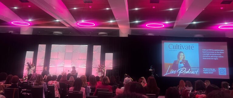 Conference room at the CNY Women's Summit in Syracuse, New York, with an audience watching a live podcast recording on stage. The lighting is pink.