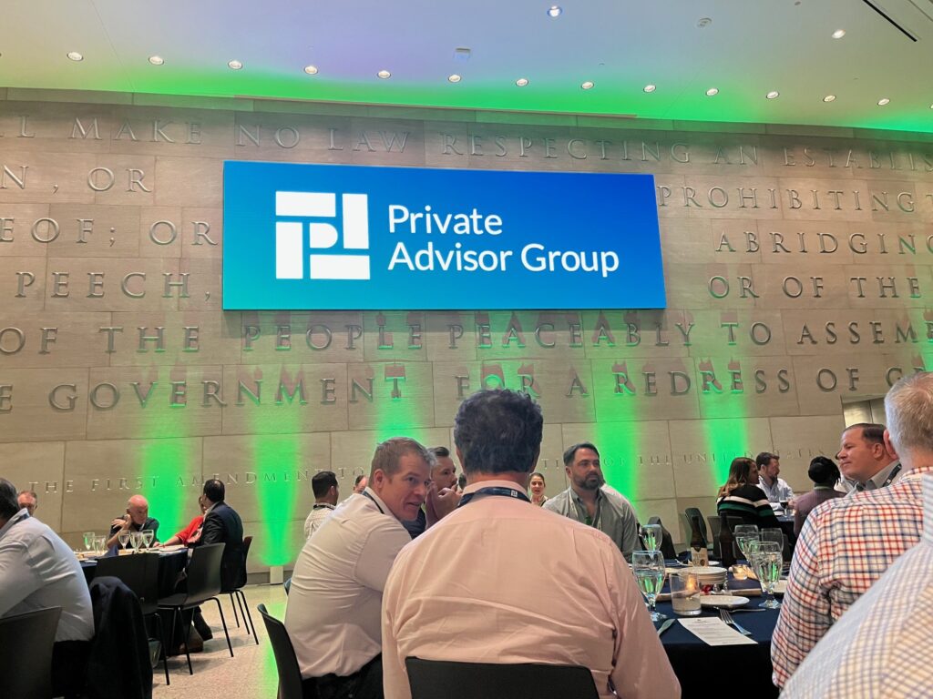 Tables with people seated for dinner inside the National Constitution Center in Philadelphia, with a large blue and white screen on the wall displaying the "Private Advisor Group" name and logo.
