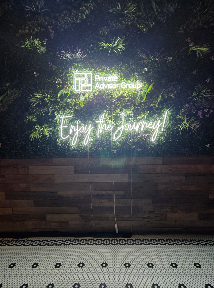 A neon white Private Advisor Group logo with the phrase "Enjoy the Journey" below it, mounted on a green indoor grass wall.