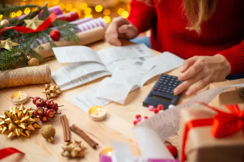 A woman sits calculating her expenses, holding receipts and using a calculator, with Christmas decorations surrounding her.