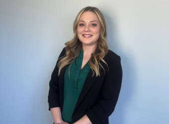woman with blonde hair and a green shirt with a black blazer standing in front of a white wall